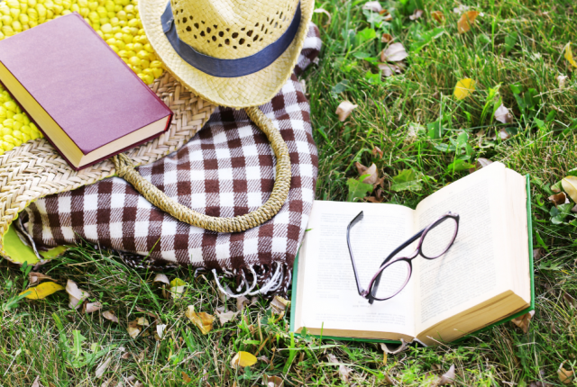 yellow straw beach bag with eyeglasses, books, a picnic blanket and a hat, spread out on a green lawn