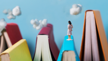 A miniature figurine of a dark haired woman stands on a mountain made of a blue book, spine side up. Additional colorful book mountains surround her. In the background is a blue sky with clouds made of cotton balls.